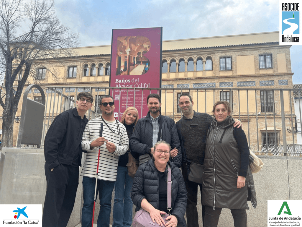 Un grupo de siete personas, personas sordociegas, guías intérpretes y voluntarios, posa sonriente frente a un cartel informativo de los Baños del Alcázar Califal. Al fondo, se observa un edificio de estilo andalusí con ventanas enrejadas y decoraciones de azulejos, rodeado por una valla metálica. El cielo está parcialmente nublado, pero la luz del día ilumina la escena.