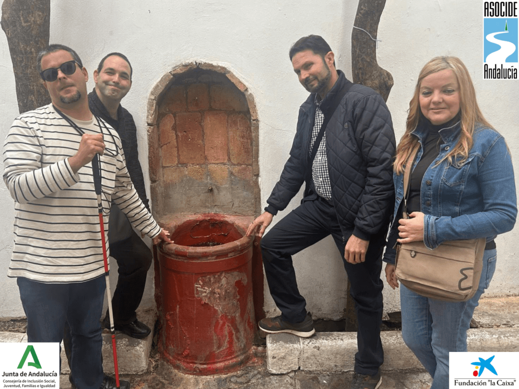 Cuatro personas sordociegas posan junto a una antigua fuente de pared con una hornacina de ladrillo y una pila cilíndrica roja desgastada. La pared blanca de fondo contrasta con la estructura de la fuente, ya los lados hay dos troncos de árboles. Nuestro socio, Sergio, está en primer plano, con su bastón rojo y blanco, mientras Rodrigo, Paco y su mujer se sitúan junto a la fuente, sonriendo. 