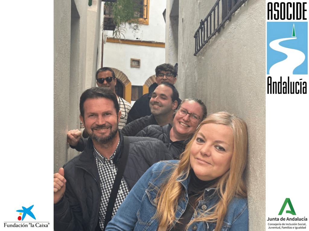 Todos los participantes posan sonrientes en un callejón estrecho de paredes blancas, acomodándose en fila a lo largo del pasillo, creando una imagen dinámica y cercana. En el fondo, se ven detalles arquitectónicos típicos andaluces, como arcos de ladrillo, ventanas con rejas y macetas con plantas colgantes.