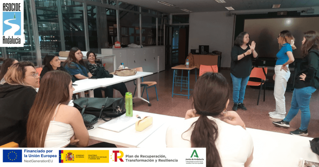 Una mujer sordociega, Mabel, vestida de negro y con jeans, se comunica mediante lengua de signos con dos alumnas de pie frente a ella. Otras estudiantes están sentadas en las mesas, observando con atención. En la imagen aparecen los logotipos de ASOCIDE Andalucía, la Unión Europea, el Gobierno de España, la Junta de Andalucía y el Plan de Recuperación, Transformación y Resiliencia."